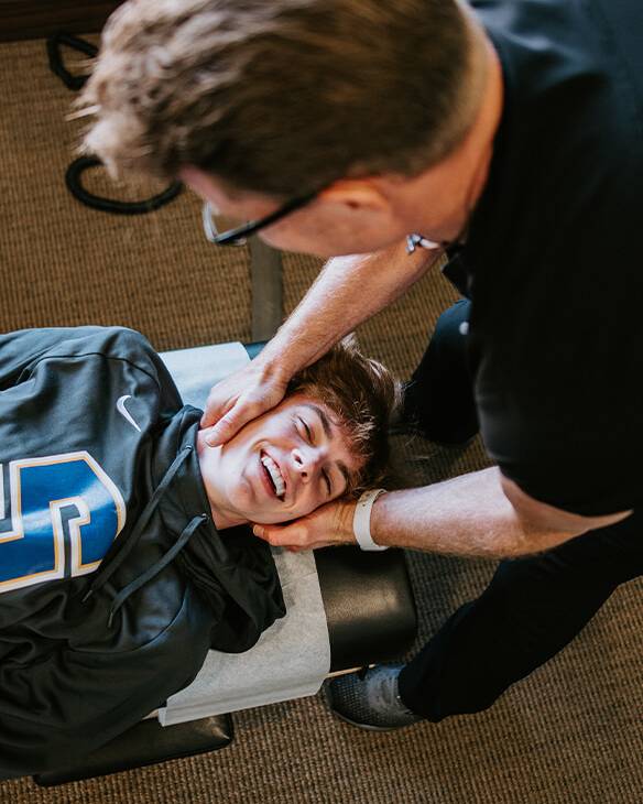 looking down from above as a chiropractor bends over to adjust a patient wearing a Stillwater sweatshirt